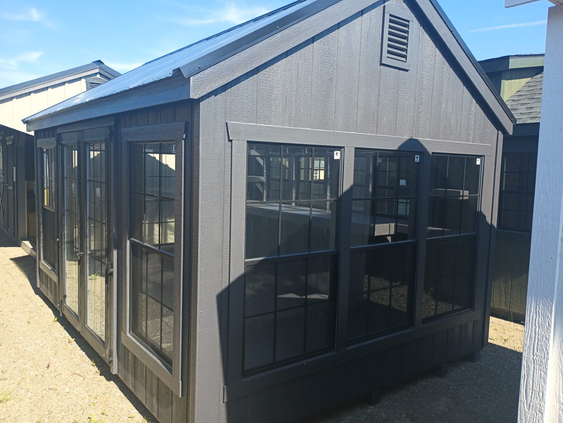 Dark color greenhouse with window surrounds, clear roof panels and shelving and a workbench on the back wall